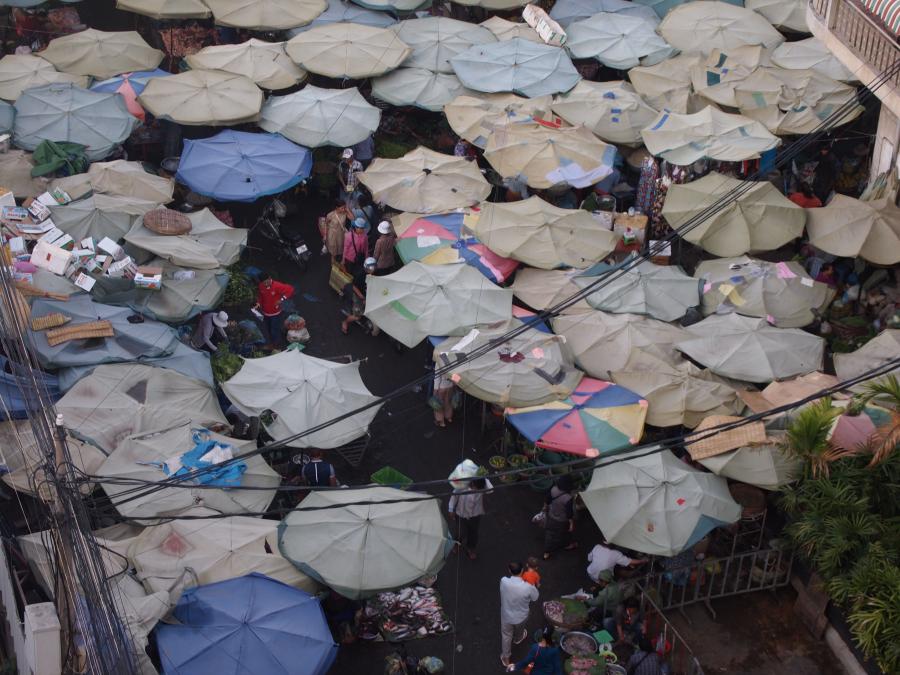 A view looking down into a street market in Phnom Penh Cambodia.  People are visible below a patchwork of round sun shades in faded blues and pinks