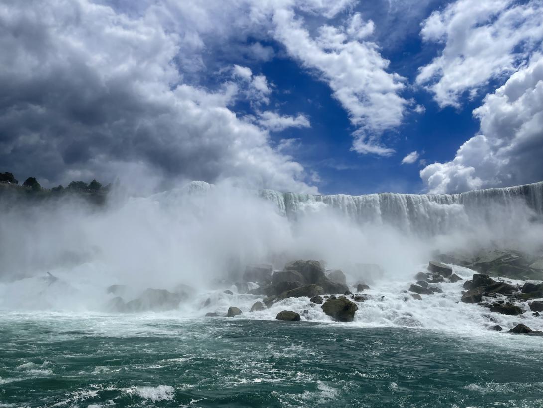 Wide photo of the US side of Niagara Falls taken from the water. Blue sky, clouds, the waterfall, rocks and blue-green water visible